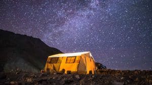 Ein großes, beleuchtetes Zelt steht auf einem felsigen Gelände unter einem atemberaubenden Nachthimmel voller Sterne. Im Hintergrund sind dunkle Bergsilhouetten zu erkennen, und ein Fahrzeug parkt neben dem Zelt. Die Szene vermittelt eine Atmosphäre von Abenteuer und Abgeschiedenheit in der Natur.