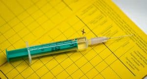 Auf dem Foto liegt eine grüne Einmalspritze mit Flüssigkeit auf einem gelben Impfpass, in dem ein Kreuz zu sehen ist.