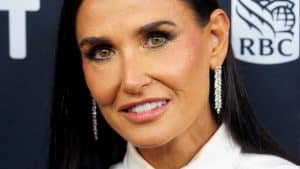 Porträt von Demi Moore, die ein weißes Hemd und eine schwarze Krawatte trägt. Sie lächelt leicht und trägt auffällige Ohrringe, während sie bei einem Event posiert.