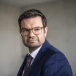 Marco Buschmann, designierter FDP-Generalsekretär, im Porträt. Er trägt einen dunkelblauen Anzug, ein weißes Hemd und eine gemusterte Krawatte, dazu eine Brille. Sein Blick ist nachdenklich, im Hintergrund ist eine schlichte Wand unscharf zu erkennen.