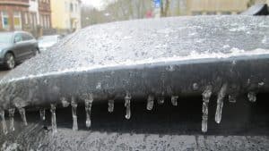 Ein Auto mit einer dünnen Eisschicht und kleinen Eiszapfen an der Heckklappe, geparkt auf einer Straße in einem Wohngebiet. Im Hintergrund sind Gebäude, Bäume und weitere parkende Autos zu sehen.