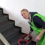 Conni in grüner Arbeitskleidung reinigt ein Treppenhaus. Sie steht neben einem Wischeimer mit Mop und schaut in die Kamera. Im Hintergrund ist eine Treppe mit dunklen Stufen zu sehen.