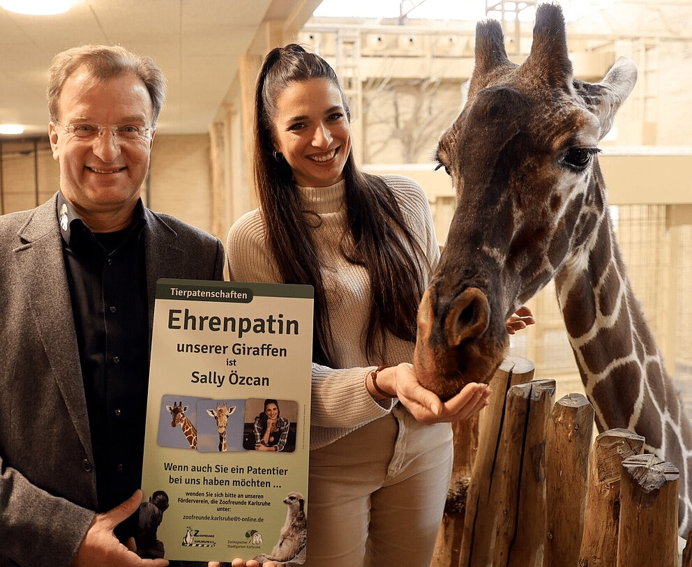 Youtuberin hautnah bei Giraffen: Sally Özcan wird Ehrenpatin im Zoo Karlsruhe 1 Zoodirektor Dr. Matthias Reinschmidt und Sally Özcan stehen vor einer Netzgiraffe im Zoo Karlsruhe und halten ein Schild, das die Ehrenpatenschaft von Sally Özcan für die Giraffen bekannt gibt.