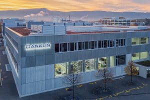 Illustration des ANKLIN-Firmengebäudes in Reinach, Schweiz, bei Sonnenuntergang. Das Gebäude zeigt eine moderne Architektur mit großen Fenstern und einem beleuchteten Firmenlogo an der Fassade. Im Vordergrund sind markierte Parkplätze für Besucher zu sehen, während im Hintergrund eine bergige Landschaft und andere Gewerbegebäude dargestellt sind.