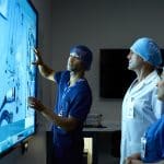 Ein Arzt im OP-Kittel erklärt auf einem großen, interaktiven Touchscreen medizinische Instrumente und Daten. Zwei weitere medizinische Fachkräfte hören aufmerksam zu. Die Szene spielt in einem modernen, technisch ausgestatteten Raum.