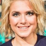 Jeanette Biedermann lächelnd vor einem bunten Hintergrund. Sie trägt ein dunkelblaues Spitzenkleid und eine dezente Halskette. Ihr blondes, welliges Haar und das elegante Make-up unterstreichen ihren glamourösen Auftritt.