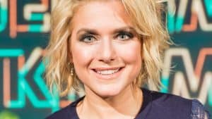 Jeanette Biedermann lächelnd vor einem bunten Hintergrund. Sie trägt ein dunkelblaues Spitzenkleid und eine dezente Halskette. Ihr blondes, welliges Haar und das elegante Make-up unterstreichen ihren glamourösen Auftritt.