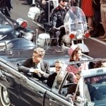 Historische Aufnahme des Präsidenten John F. Kennedy in einer offenen Limousine während einer öffentlichen Parade, begleitet von First Lady Jacqueline Kennedy. Sicherheitskräfte und Zuschauer säumen die Straßen.