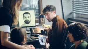 Swen sitzt nachdenklich an einem Schreibtisch, während ein Ermittlerteam um ihn herum arbeitet. Auf einem Monitor im Hintergrund ist das Bild eines Mannes zu sehen, und in einer Hand wird ein Foto gezeigt. Die Szene zeigt eine angespannte Atmosphäre, während die Ermittler auf Swens Erinnerungen hoffen, um den Fall zu lösen.