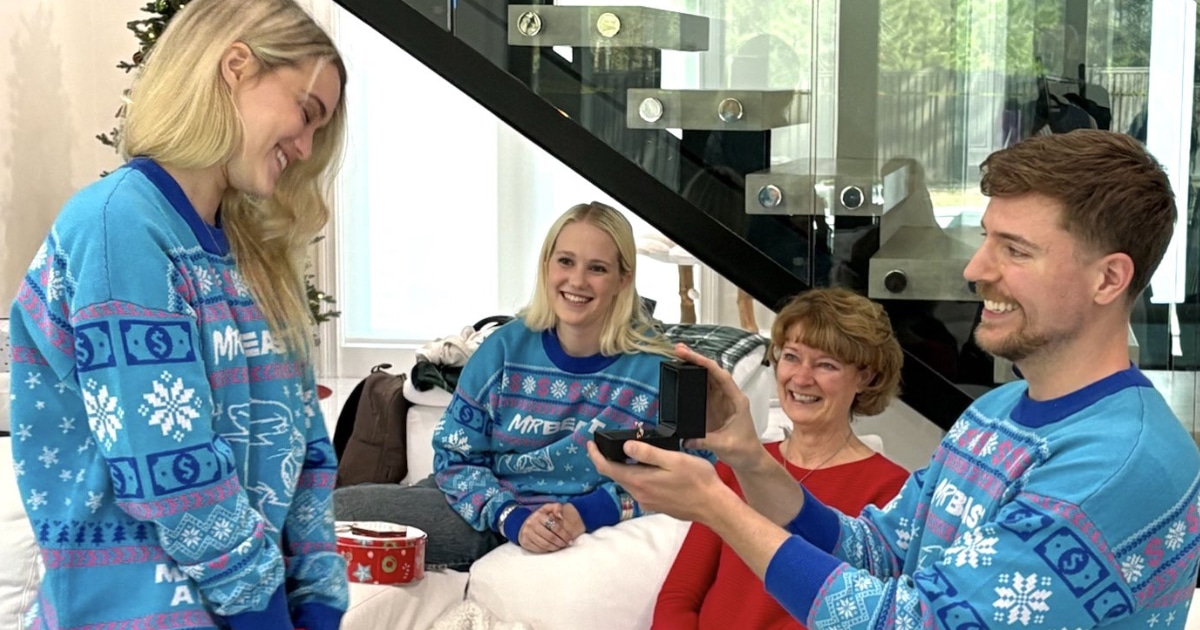 MrBeast macht seiner Freundin Thea einen Heiratsantrag. Er kniet auf einem weißen Teppich und präsentiert eine Ringbox, während Thea lächelt. Im Hintergrund sitzen zwei Frauen, darunter vermutlich Familienmitglieder, die den Moment freudig beobachten.