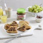 Ein gedeckter Frühstückstisch mit belgischen Waffeln, die mit nutella Plant-Based bestrichen sind. Im Hintergrund steht ein Glas nutella Plant-Based mit grünem Deckel, umgeben von Trauben, einem Glas Orangensaft und weiteren Frühstückszutaten.