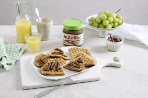 Ein gedeckter Frühstückstisch mit belgischen Waffeln, die mit nutella Plant-Based bestrichen sind. Im Hintergrund steht ein Glas nutella Plant-Based mit grünem Deckel, umgeben von Trauben, einem Glas Orangensaft und weiteren Frühstückszutaten.