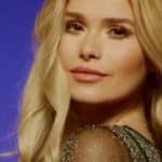 Pamela Reif in einem eleganten, schwarzen Kleid mit glitzernden Ärmeln. Ihre langen blonden Haare fallen sanft über die Schultern, während sie in einem stimmungsvoll beleuchteten Studio mit violettem Hintergrund in die Kamera schaut.