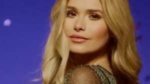 Pamela Reif in einem eleganten, schwarzen Kleid mit glitzernden Ärmeln. Ihre langen blonden Haare fallen sanft über die Schultern, während sie in einem stimmungsvoll beleuchteten Studio mit violettem Hintergrund in die Kamera schaut.