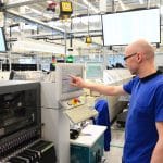 Ein Mitarbeiter in einem modernen Produktionswerk bedient ein Touchscreen-Panel an einer Fertigungsmaschine. Im Hintergrund sind weitere Arbeitsplätze, technische Geräte und Monitore mit Produktionsdaten zu sehen. Die Umgebung ist hell und organisiert, mit industriellen Anlagen und Bildschirmen, die die Effizienz der Produktionsprozesse unterstützen.