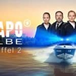 Alt-Text: "Cover von 'WaPo Elbe, Staffel 2' zeigt das Ermittlerteam auf einem Polizeiboot, das auf dem Fluss patrouilliert, mit dramatischem Sonnenuntergang im Hintergrund."