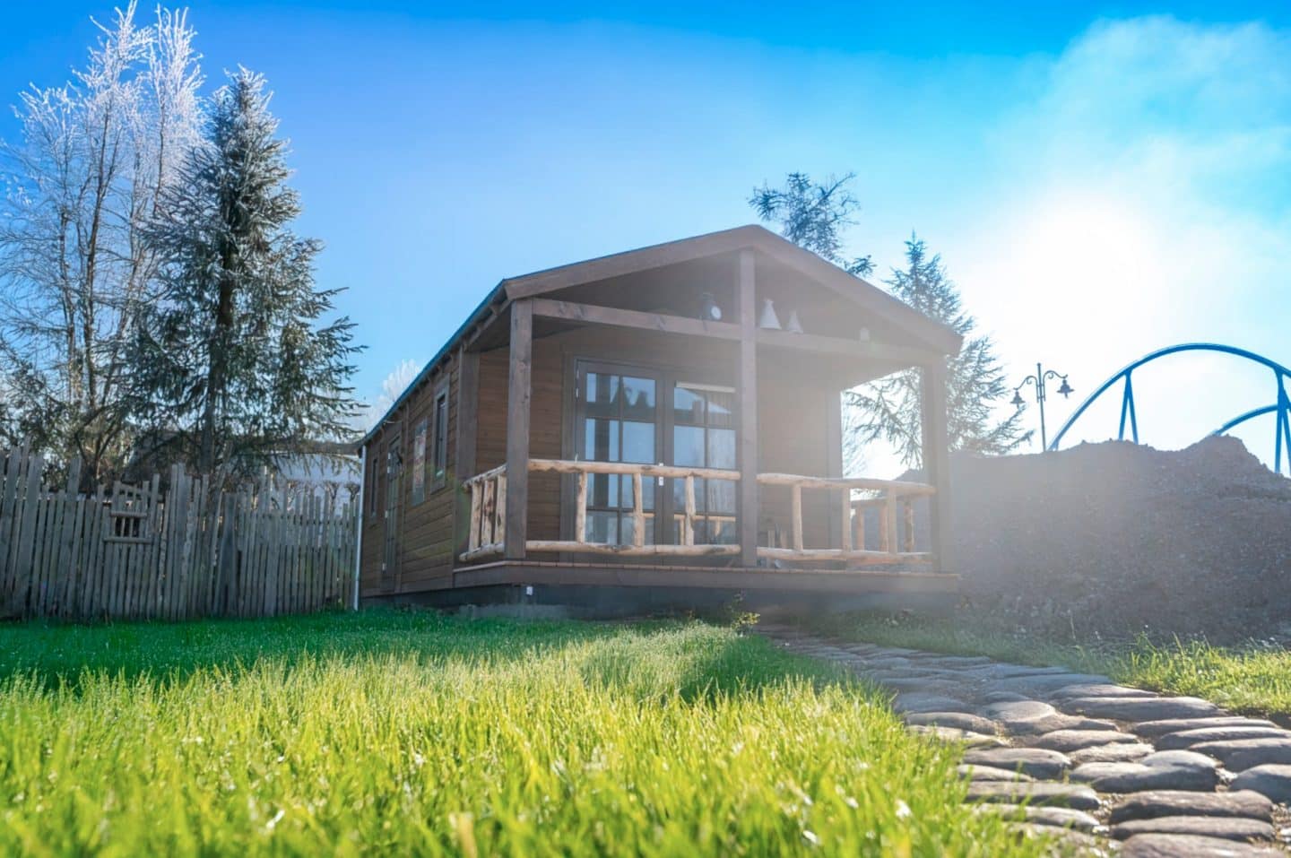 Europa-Park plant riesige Westernstadt „Silver Lake City“ – Eröffnung bis 2026Neues Western-Erlebnis in Rust 1 Eine rustikale Blockhütte mit überdachter Veranda im Westernstil, umgeben von grüner Wiese und Holzzaun, mit einer Achterbahnstruktur im Hintergrund unter blauem Himmel.