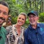 Die Moderatoren Jan Köppen und Sonja Zietlow zusammen mit Dr. Bob im Dschungelcamp. Sie lächeln in die Kamera, während im Hintergrund der typische Dschungel-Set-Aufbau mit Büchern und Kisten zu sehen ist.