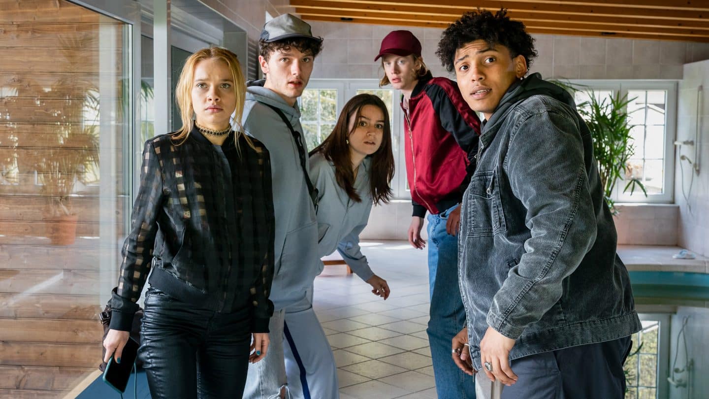 "Tatort: Herz der Dunkelheit" – Tödliche Party-Nacht! Gorniaks letzter Fall wird zum Albtraum 3 Fünf Jugendliche stehen angespannt in einem modernen Poolhaus mit Glaswänden und Fliesenboden. Maya hält ein Handy in der Hand, während alle mit erschrockenen und überraschten Blicken in eine Richtung schauen. Ihre Körperhaltung wirkt angespannt, als wären sie gerade ertappt worden