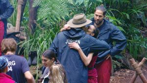 Jürgen Hingsen verabschiedet sich im Dschungelcamp von Lilly Becker mit einer innigen Umarmung. Maurice Dziwak steht daneben und beobachtet die Szene. Im Hintergrund sind weitere Camp-Bewohner sowie tropische Vegetation zu sehen.