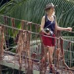 Nina Bott verlässt das Dschungelcamp und läuft über die ikonische Hängebrücke, umgeben von tropischer Vegetation. Sie trägt einen Hut, ein dunkelblaues Top und rote Shorts, während sie mit einem Lächeln in die Ferne blickt. In ihrer Hand hält sie einen kleinen Gegenstand.