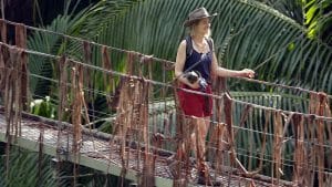 Nina Bott verlässt das Dschungelcamp und läuft über die ikonische Hängebrücke, umgeben von tropischer Vegetation. Sie trägt einen Hut, ein dunkelblaues Top und rote Shorts, während sie mit einem Lächeln in die Ferne blickt. In ihrer Hand hält sie einen kleinen Gegenstand.