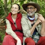 Lilly Becker und Pierre Sanoussi-Bliss sitzen im Dschungelcamp. Lilly Becker trägt eine rote Weste und lächelt, während Pierre Sanoussi-Bliss mit Hut und Brille entspannt neben ihr sitzt und eine Feldflasche hält. Beide wirken zufrieden.