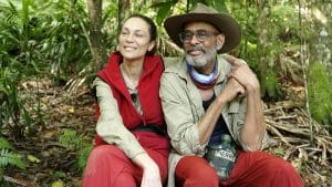 Lilly Becker und Pierre Sanoussi-Bliss sitzen im Dschungelcamp. Lilly Becker trägt eine rote Weste und lächelt, während Pierre Sanoussi-Bliss mit Hut und Brille entspannt neben ihr sitzt und eine Feldflasche hält. Beide wirken zufrieden.