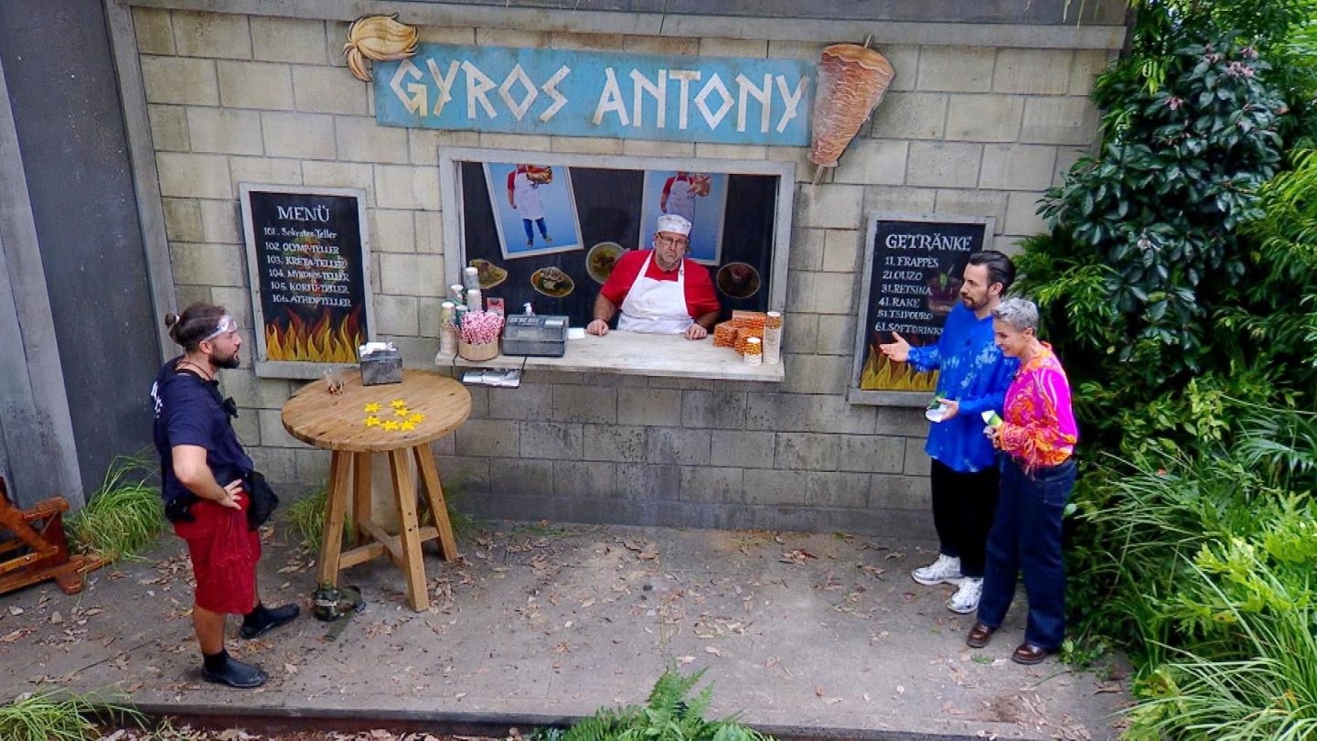 Maurice Dziwak steht nach der Dschungelprüfung „Einkaufs-Strafe“ vor einem Imbissstand namens „Gyros Antony“. Er trägt Schutzbrille und rote Shorts, während Sonja Zietlow und Jan Köppen ihn beglückwünschen. Auf dem Tisch liegen sieben von zehn gesammelten Sternen. Im Hintergrund ist ein Verkäufer in roter Schürze zu sehen, der hinter dem Tresen steht. Die Szene spielt sich inmitten einer künstlichen Dschungelumgebung ab.







