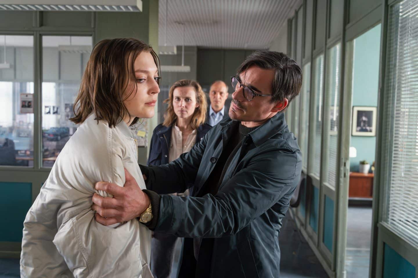 "Tatort: Herz der Dunkelheit" – Tödliche Party-Nacht! Gorniaks letzter Fall wird zum Albtraum 4 Romy Brahms steht mit ernster Miene im Verhörraum, während ihr Vater Paul Brahms sie fest an den Schultern hält und eindringlich ansieht. Hinter ihnen beobachten Karin Gorniak und Peter Michael Schnabel das Geschehen durch eine Glaswand. Die Stimmung im Raum ist angespannt.