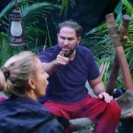 Maurice Dziwak sitzt wütend im Dschungelcamp und zeigt mit dem Finger auf Anna-Carina Woitschack, die ihm den Rücken zuwendet. Seine Gesichtszüge sind angespannt, er scheint lautstark zu sprechen. Links im Bild beobachtet Alessia Herren die Szene mit ernster Miene. Die grüne Dschungelkulisse und eine Laterne im Hintergrund verstärken die dramatische Atmosphäre der hitzigen Diskussion.