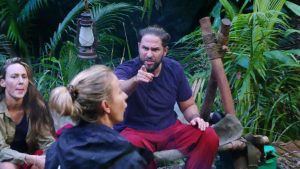 Maurice Dziwak sitzt wütend im Dschungelcamp und zeigt mit dem Finger auf Anna-Carina Woitschack, die ihm den Rücken zuwendet. Seine Gesichtszüge sind angespannt, er scheint lautstark zu sprechen. Links im Bild beobachtet Alessia Herren die Szene mit ernster Miene. Die grüne Dschungelkulisse und eine Laterne im Hintergrund verstärken die dramatische Atmosphäre der hitzigen Diskussion.