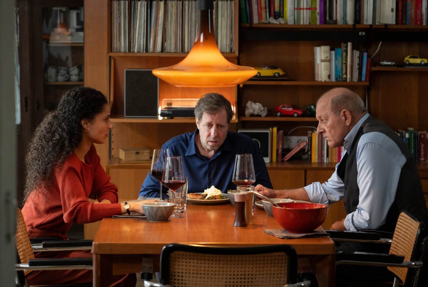 "Wilsberg - Achtsam bis tödlich": In einer Wohnung sitzen Wilsberg (Leonard Lansink), Ekki Talkötter (Oliver Korittke) und Tessa Tilker (Patricia Meeden) um einen gedeckten Tisch herum. Sie schauen sich ernst an. Vor ihnen stehen Teller mit Essen und Weingläser.
