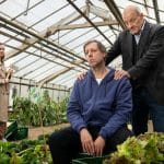 "Wilsberg - Achtsam bis tödlich": Ekki Talkötter (Oliver Korittke) sitzt in einem Gewächshaus, in welchem Gemüse angepflanzt wird, unmotiviert auf einer Kiste. Wilsberg (Leonard Lansink) steht hinter Ekki, legt ihm die Hände väterlich auf die Schultern und schaut ihn besorgt an. Im Hintergrund steht Silke Sestendrup (Nadja Becker) mit einer Axt in der Hand und lächelt.
