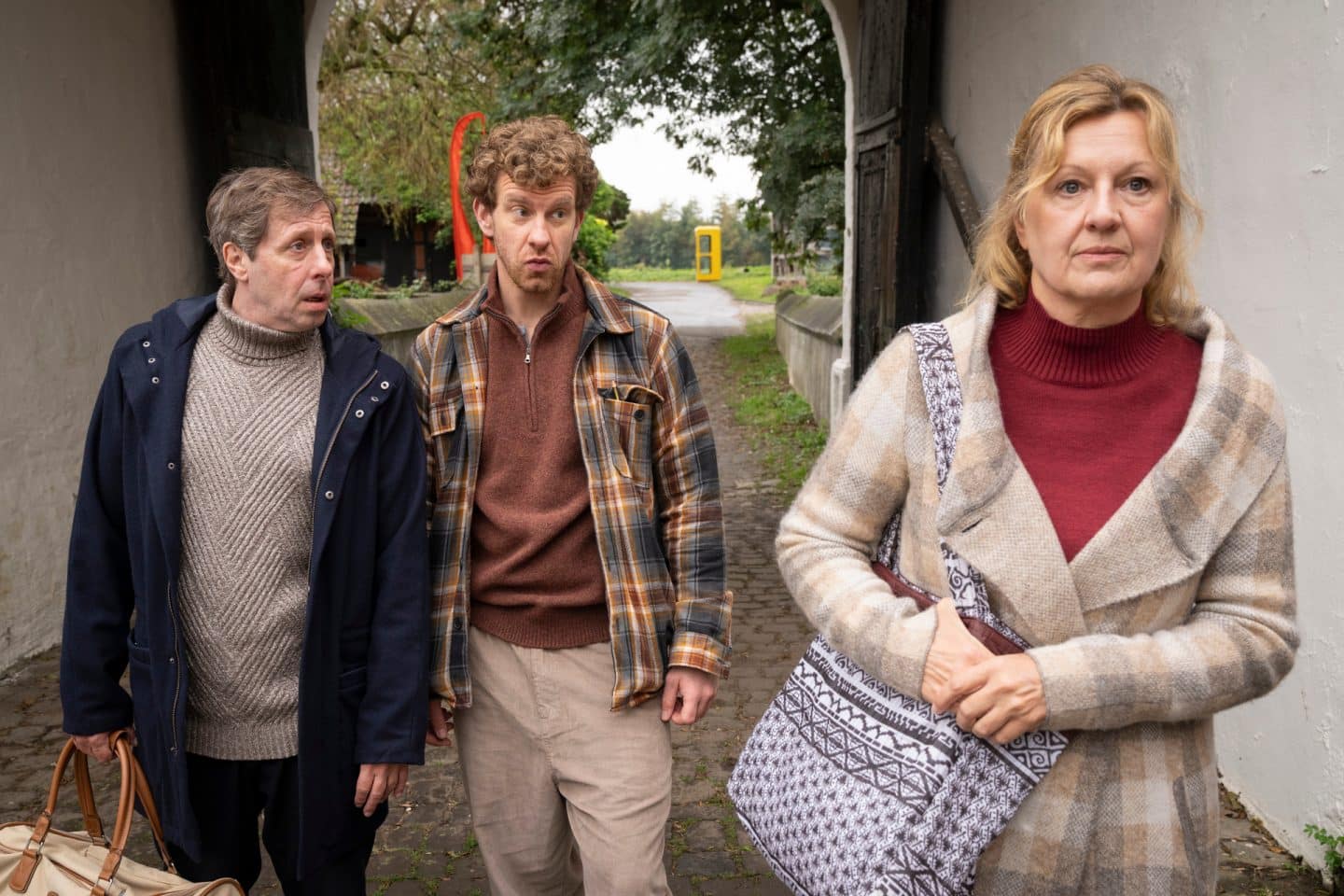 "Wilsberg - Achtsam bis tödlich": Ekki Talkötter (Oliver Korittke) läuft neben Mikka Lindental (Sebastian Weiss) durch einen Torbogen hindurch, Renate Trimborn (Claudia Lietz) eilt an ihnen vorbei und hält ihre Handtasche dabei fest, die sie über ihrer Schulter trägt.
