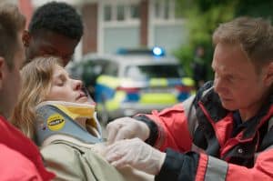 "Notruf Hafenkante - Kein Zurück": Großaufnahme von Dr. Haase (Fabian Harloff) und Julia Jacobi (Inga Lessmann). Dr. Haase versorgt Julia Jacobi, die auf einer Patiententrage liegt, daneben stehen zwei Sanitäter (Komparsen). Im Hintergrund steht ein Streifenwagen.