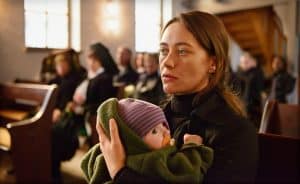 "Spreewaldkrimi - Böses muss mit Bösem enden": Fina Jurisch (Mercedes Müller) sitzt in einer Kirche und hat ihr Baby auf ihrem Schoß. Im Hintergrund sitzen weitere Personen (Komparsen) in Kirchenbänken.