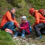 "Der Bergdoktor: Hoffnung": An einem begrünten Berghang: Tamara Gotthart (Rosa Wirtz) sitzt mit schmerzverzerrtem Gesicht am Boden. Sie trägt einen Helm mit Stirnlampe und einen Rucksack. Neben ihr sitzt Dr. Martin Gruber (Hans Sigl) und tastet ihren Arm ab, auf der anderen Seite kniet Hans Gruber (Heiko Ruprecht), der seine Bergretter-Ausrüstung trägt.
