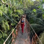 Yeliz Koc läuft über eine schmale Hängebrücke inmitten eines dichten, tropischen Regenwaldes. Sie trägt einen Cowboyhut, ein schwarzes Top mit Aufdruck und eine rote Hose. Umgeben von üppigem Grün und hohen Palmen, blickt sie konzentriert nach vorne.