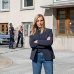"Die Jägerin - Gegen die Wut": Staatsanwältin Judith Schrader (Nadja Uhl) steht mit verschränkten Armen vor einer Polizeidienststelle. Hinter ihr sind eine Polizistin (Komparsin) und ein Polizist (Komparse) in Uniform im Gespräch..