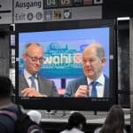"Kanzler und Herausforderer - Scholz und Merz im Wahlkampf": Fotomontage: Auf einem Bildschirm in einer U-Bahnstation sind Friedrich Merz und Olaf Scholz zu sehen. Im Vordergrund sind Hinterköpfe von Menschen, im Hintergrund über dem Bildschirm befinden sich Wegweiser.