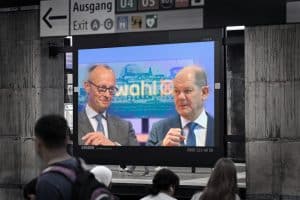 "Kanzler und Herausforderer - Scholz und Merz im Wahlkampf": Fotomontage: Auf einem Bildschirm in einer U-Bahnstation sind Friedrich Merz und Olaf Scholz zu sehen. Im Vordergrund sind Hinterköpfe von Menschen, im Hintergrund über dem Bildschirm befinden sich Wegweiser.