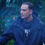Timur Ülker steht mit ernster Miene und ausgebreiteten Armen im Dschungelcamp, während er in einer hitzigen Diskussion spricht. Er trägt eine dunkle Regenjacke, sein Gesichtsausdruck zeigt Entschlossenheit und Emotion. Der grüne Dschungelhintergrund mit Laterne und Pflanzen verstärkt die dramatische Atmosphäre des Moments, in dem er am Lagerfeuer eine Debatte entfacht.