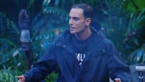 Timur Ülker steht mit ernster Miene und ausgebreiteten Armen im Dschungelcamp, während er in einer hitzigen Diskussion spricht. Er trägt eine dunkle Regenjacke, sein Gesichtsausdruck zeigt Entschlossenheit und Emotion. Der grüne Dschungelhintergrund mit Laterne und Pflanzen verstärkt die dramatische Atmosphäre des Moments, in dem er am Lagerfeuer eine Debatte entfacht.