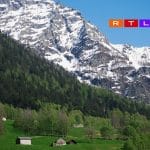 "Landschaft mit grünen Wiesen und Holzhütten vor schneebedeckten Bergen. Im oberen rechten Bereich sind die Logos von RTL+ und SRF zu sehen."