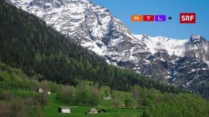 "Landschaft mit grünen Wiesen und Holzhütten vor schneebedeckten Bergen. Im oberen rechten Bereich sind die Logos von RTL+ und SRF zu sehen."