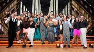 Gruppenfoto der 'Let's Dance'-Kandidaten auf der Bühne. Die Promis und Profis posieren gemeinsam in eleganten Outfits, viele lächeln und zeigen jubelnde Gesten. Im Hintergrund ist das festlich beleuchtete Show-Set zu sehen.