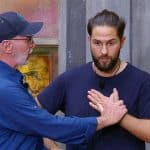 Maurice Dziwak steht mit ernster Miene vor einer grauen Wand, während Dr. Bob ihm Anweisungen gibt. Der Mediziner trägt eine blaue Jacke und eine Kappe, legt eine Hand auf Maurices Brust und scheint ihm etwas zu erklären. Die Szene spielt sich im Rahmen der Dschungelprüfung „Einkaufs-Strafe“ ab.