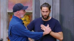 Maurice Dziwak steht mit ernster Miene vor einer grauen Wand, während Dr. Bob ihm Anweisungen gibt. Der Mediziner trägt eine blaue Jacke und eine Kappe, legt eine Hand auf Maurices Brust und scheint ihm etwas zu erklären. Die Szene spielt sich im Rahmen der Dschungelprüfung „Einkaufs-Strafe“ ab.