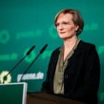 Franziska Brantner steht an einem Rednerpult vor einer grünen Wand mit dem Logo und der Schrift der Partei Bündnis 90/Die Grünen. Sie trägt eine Brille, eine dunkle Jacke und eine olivgrüne Bluse. Vor ihr befinden sich zwei Mikrofone.